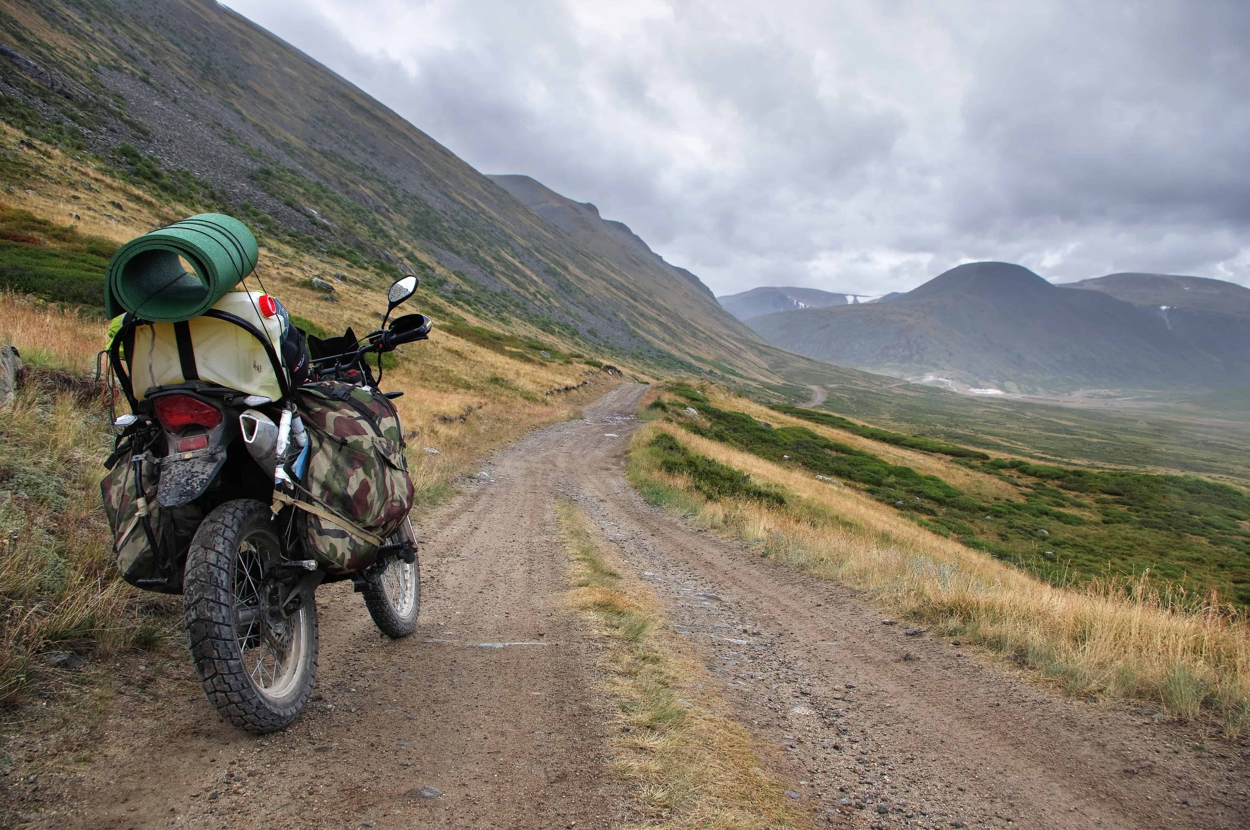 Motorcycle enduro traveler with suitcases standing on extreme rocky road in a mountain valley in cloudy weather on the background of gloom fog mountain ranges.